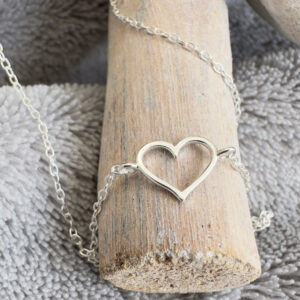 Silver anklet with a charm of an outline of a heart displayed on wood.