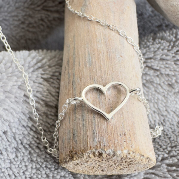 Silver anklet with a charm of an outline of a heart displayed on wood.