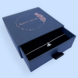 Captivating silver anklet with an evil eye charm with white gemstone in the centre, presented in a blue jewellery box with Anklet Allure written on the top in rose gold.