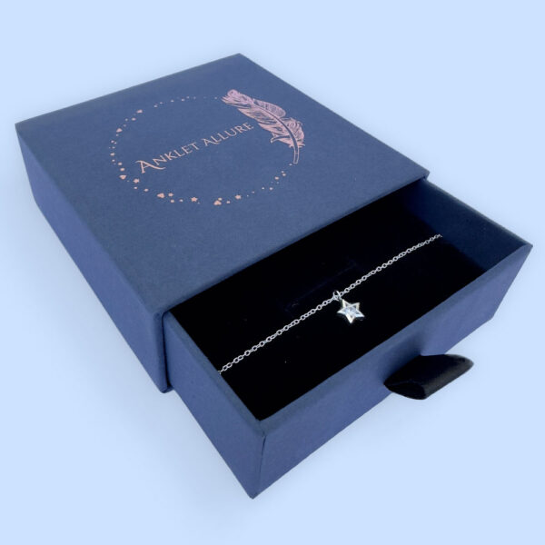 Ankle bracelet with a star charm with a white gemstone in the centre, presented in a blue jewellery box with Anklet Allure written on the top in rose gold.