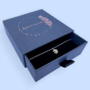 Spiritual anklet with a silver ohm symbol charm, presented in a blue jewellery box with Anklet Allure written on the top in rose gold.