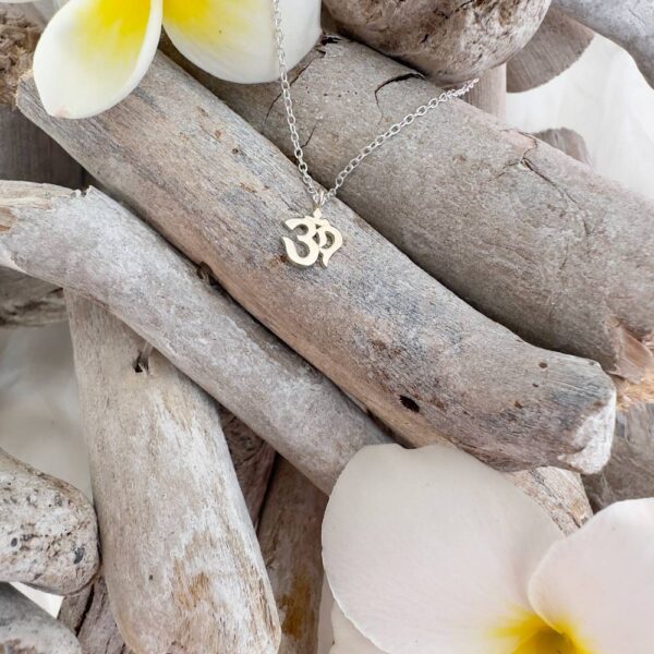 Silver anklet with an ohm symbol pendant presented on wood sticks with yellow and white frangipani flowers petal in top left and bottom right corners.