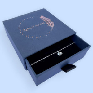 Silver anklet with a charm of a cute elephant with a long trunk, presented in a blue jewellery box with Anklet Allure written on the top in rose gold.