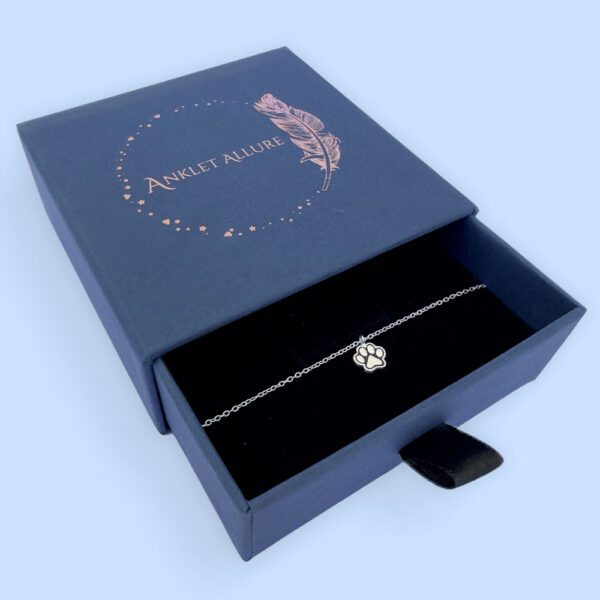 Silver anklet with a charm of a dog's pawprint with ionised silver detail, presented in a blue jewellery box with Anklet Allure written on the top in rose gold.