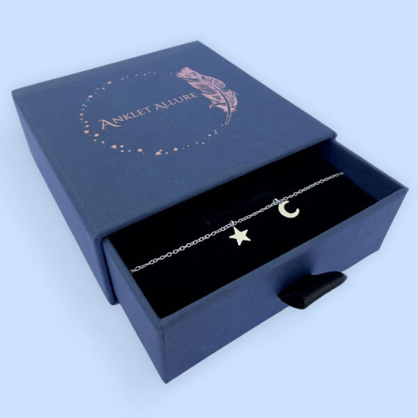 Celestial anklet with a star and a crescent moon, presented in a blue jewellery box with Anklet Allure written on the top in rose gold.