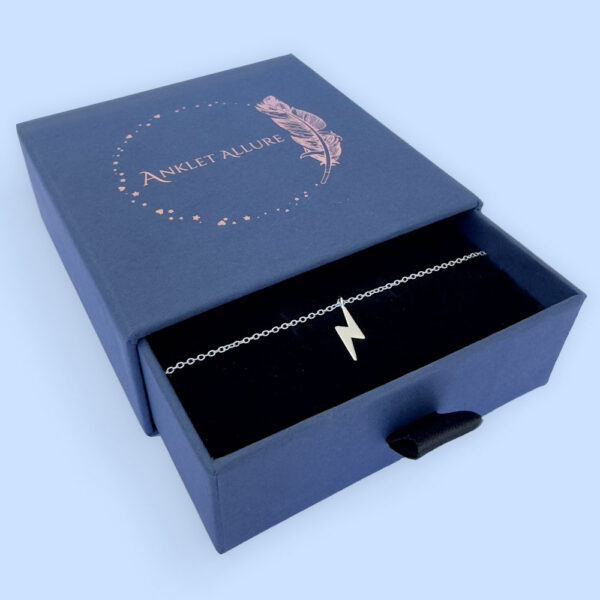 Powerful silver anklet with a charm of a lightening bolt, presented in a blue jewellery box with Anklet Allure written on the top in rose gold.