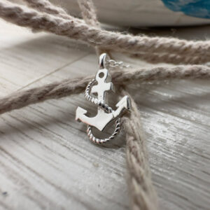 Close up of a silver anklet charm. A silver anchor with silver rope wrapped around it sitting on a rope with a white washed wood background.