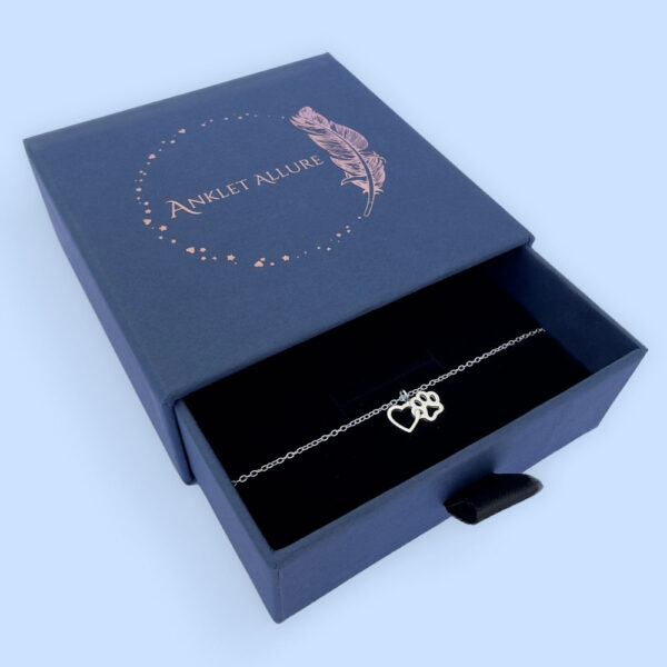 Silver anklet of a linked heart and pawprint, presented in a blue jewellery box with Anklet Allure written on the top in rose gold.