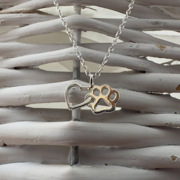 Close up of a sterling silver heart and pawprint charm interlinked together on an ankle bracelet on a white basket background.