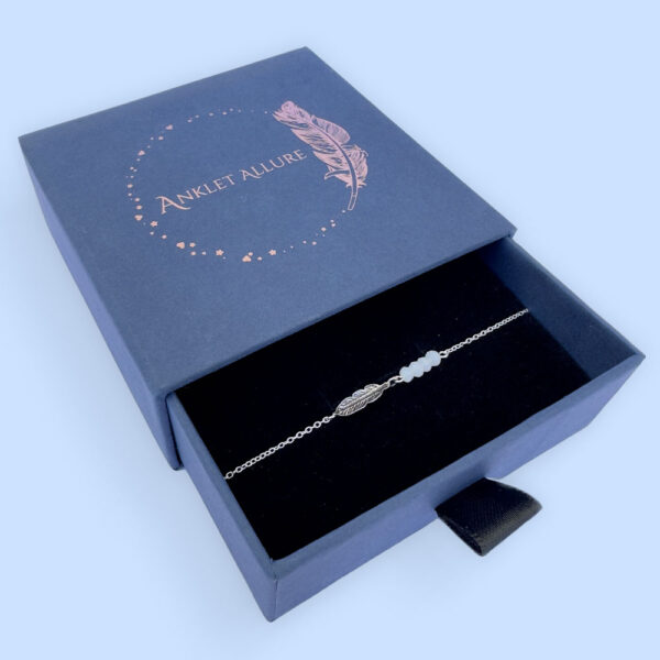 Anklet with an iodised silver style feather charm and four white beads, presented in a blue jewellery box with Anklet Allure written on the top in rose gold.