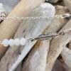 Close up of silver ankle bracelet with an oxidised silver feather charm attached to four white beads, with white-washed wood in the background.