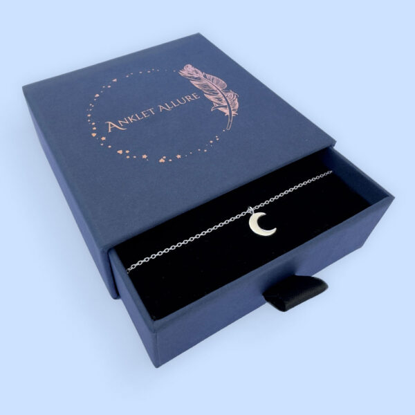 Beautiful silver anklet with a crescent moon charm, presented in a blue jewellery box with Anklet Allure written on the top in rose gold.