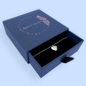Silver ankle bracelet with a large flat heart charm with a white gemstone, presented in a blue jewellery box with Anklet Allure written on the top in rose gold.