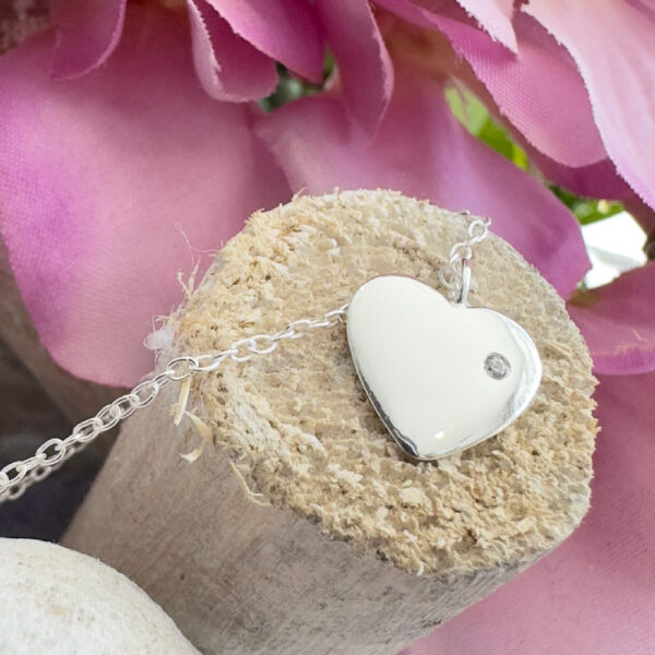 Solid sterling silver anklet with a heart charm with a dainty white gem displayed on the end of a stick with purple petals in the top right background.