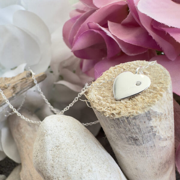 Solid sterling silver anklet with a heart charm with a dainty white gem displayed on the end of a stick with purple petals in the top right background.