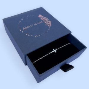 Anklet in silver with a stunning gem encrusted cross within the chain, presented in a blue jewellery box with Anklet Allure written on the top in rose gold.
