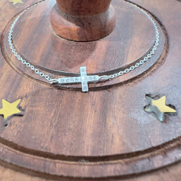 Ankle bracelet with a cross along a section of the chain with twelve white gemstones along the cross on a wooden base.