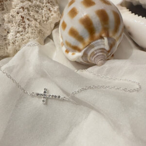 Stunning silver anklet with a cross on it and white gemstones along the cross. Displayed on a white sheet with a brown and white shell and pumice in the background.