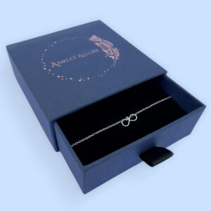 Eye catching silver anklet with a dainty infinity charm, presented in a blue jewellery box with Anklet Allure written on the top in rose gold.
