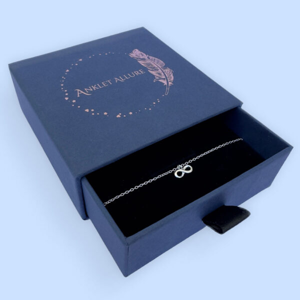 Close up of a silver anklet with an infinity charm, presented in a blue jewellery box with Anklet Allure written on the top in rose gold.