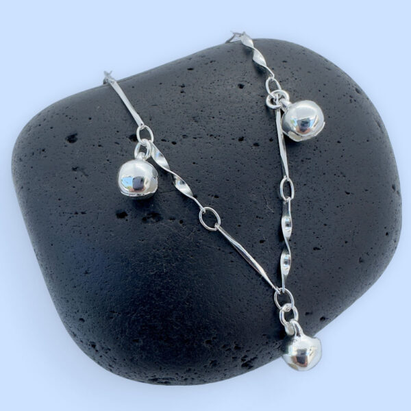 Silver anklet with a stiff twisted chain and three bells equally spaced around, presented on a black rock.