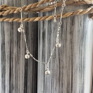 Silver anklet with jingling balls on a silver chain designed to look twisted, on a black and white painted wood background with rope around the top.