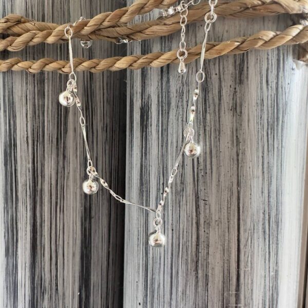 Silver anklet with jingling balls on a silver chain designed to look twisted, on a black and white painted wood background with rope around the top.