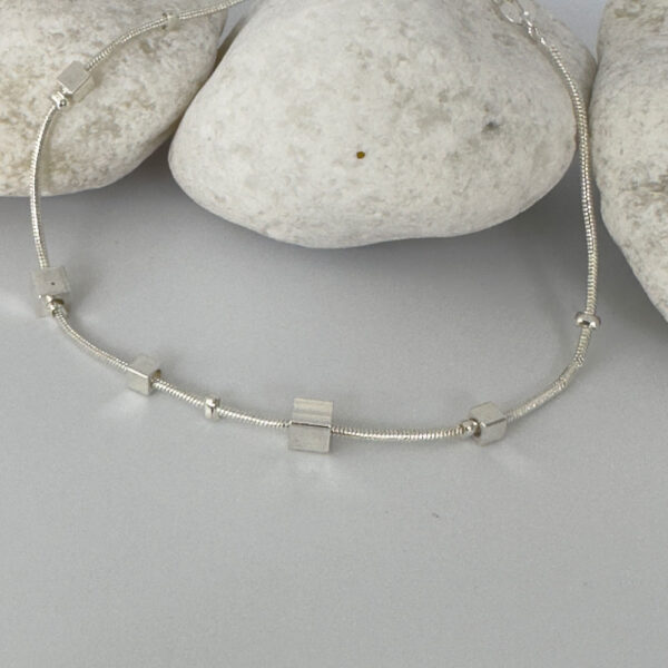 Ankle bracelet in silver with different sized square charms on white rocks.