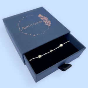 Ankle bracelet in silver with different sized square charms in a blue jewellery box.