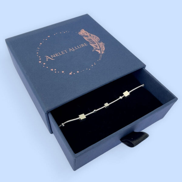 Ankle bracelet in silver with different sized square charms in a blue jewellery box.