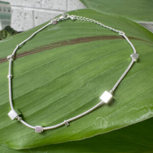Shiny silver anklet with five silver square charms around it presented on a big green leaf.