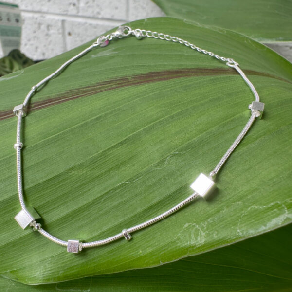 Shiny silver anklet with five silver square charms around it presented on a big green leaf.
