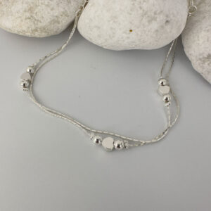 Double chain silver anklet with a heart in between two silver balls on each chain on a white rock.