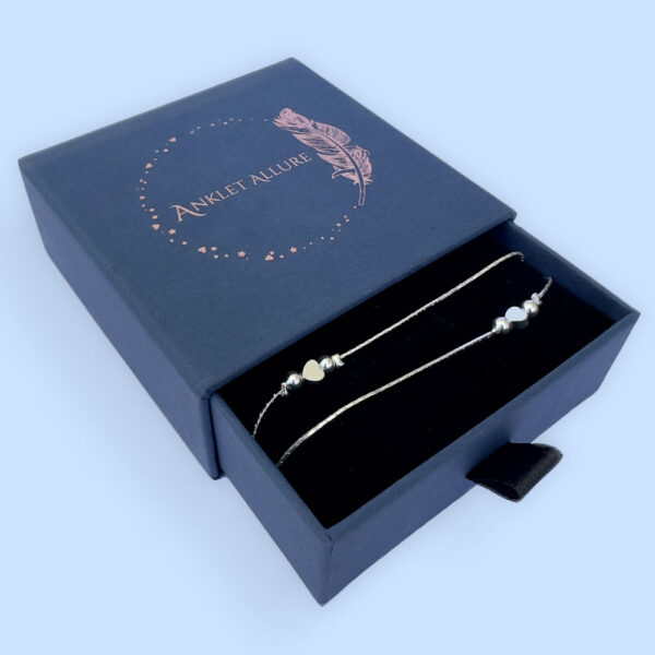 Double chain silver anklet with a heart in between two silver balls on each chain in a blue jewellery box.