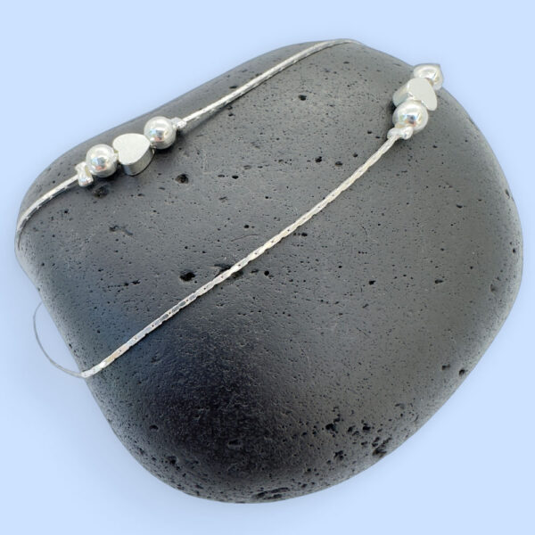 Double chain silver anklet with a heart in between two silver balls on each chain, displayed on a black rock.