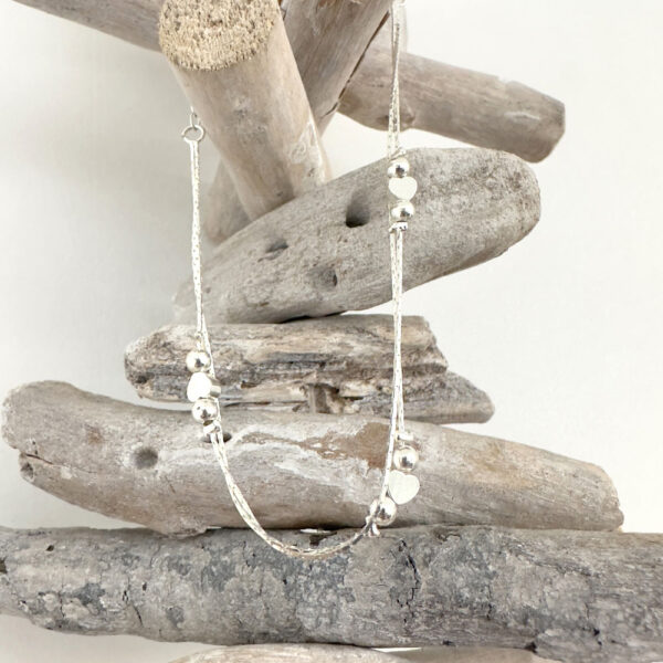 Double chain silver anklet with a heart between two silver balls, repeated three times around the chain, hanging from white washed wood pieces piled up.