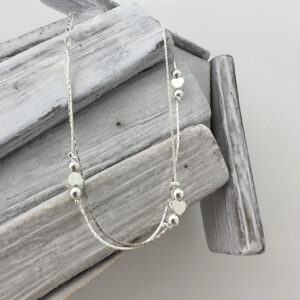 Double chain silver anklet with a heart between two silver balls, repeated three times around the chain, hanging from white washed wood