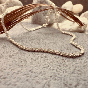A simple silver anklet that sparkles, presented on a charcoal coloured rock hanging over a brown leaf.