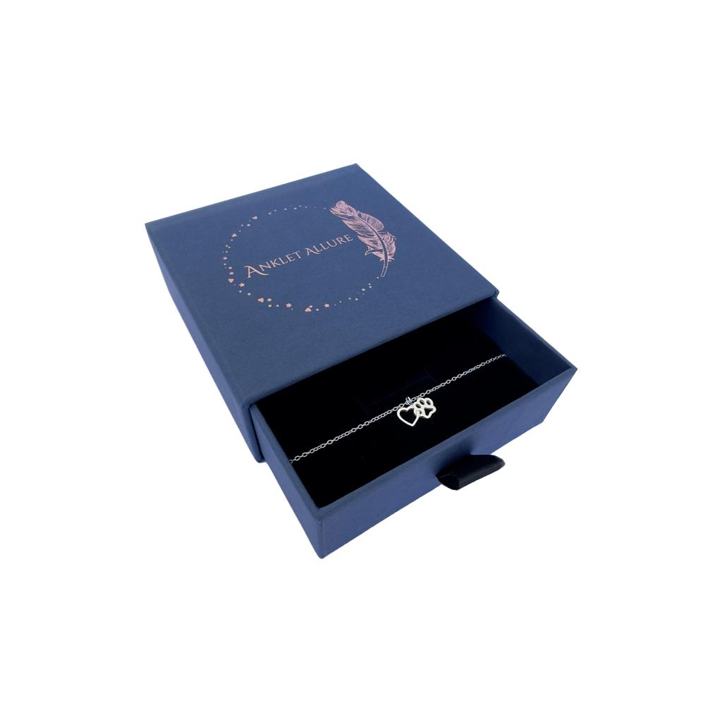 Cute anklet with interlinked pawprint and heart charm, presented in a blue jewellery box with Anklet Allure written on the top in rose gold.