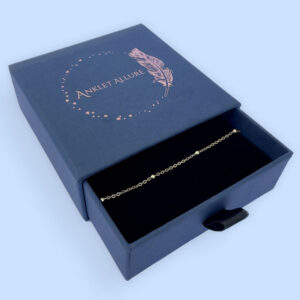 Gold chain with three small gold balls presented in a dark blue Anklet Allure gift box.