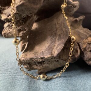Close up of gold ankle bracelet with small balls around the chain hanging from some driftwood.