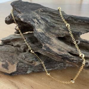 Elegant gold anklet with small gold balls spread around a gold chain at equal distance. Displayed on a piece of driftwood.