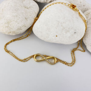 Stunning double chain gold anklet with an infinity charm with fifteen white gemstones along half of it on a white rock.