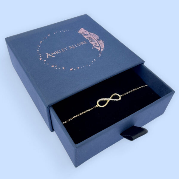 Dainty gold double chain anklet with a sold gold heart charm presented in a dark blue Anklet Allure gift box.