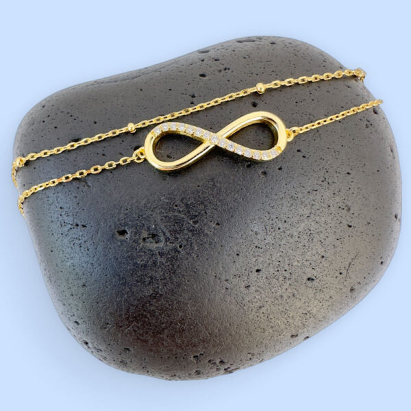 Stunning double chain gold anklet with an infinity charm with fifteen white gemstones along half of it, displayed on a black rock.