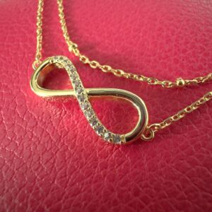 Close up of a gold infinity charm covered with cubic zirconia gems along one half, on a stunning gold ankle bracelet.