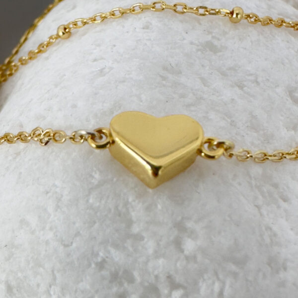 Close up of a dainty gold double chain anklet with a sold gold heart charm on a white rock.