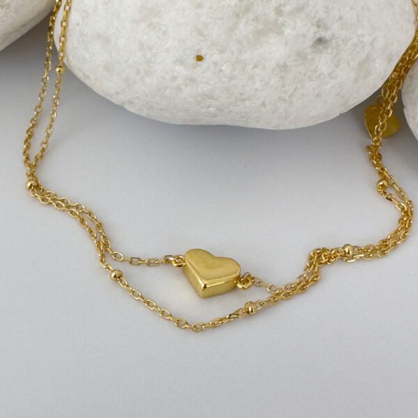 Dainty gold double chain anklet with a sold gold heart charm on a white rock.