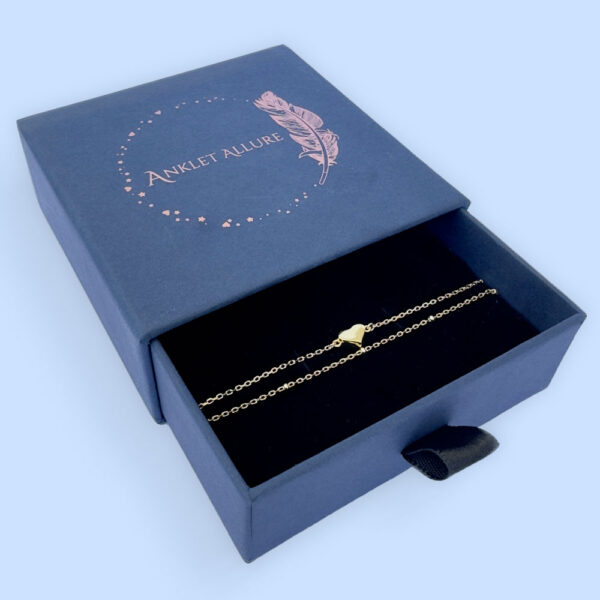 Dainty gold double chain anklet with a sold gold heart charm presented in a dark blue Anklet Allure gift box.