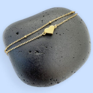Dainty gold double chain anklet with a sold gold heart charm on a black rock.